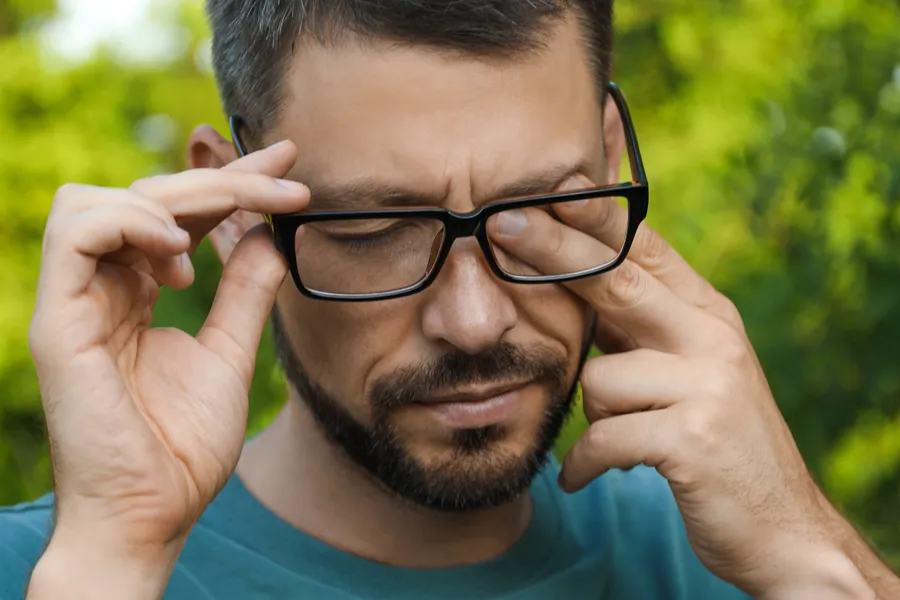 A man with a beard and glasses standing outside and wearing a blue t-shirt lifts his glasses and rubs his eyes due to allergy irritation. Get treatment for allergic conjunctivitis from Allen Green, M.D. of Center for Optimum Health in Los Angeles.