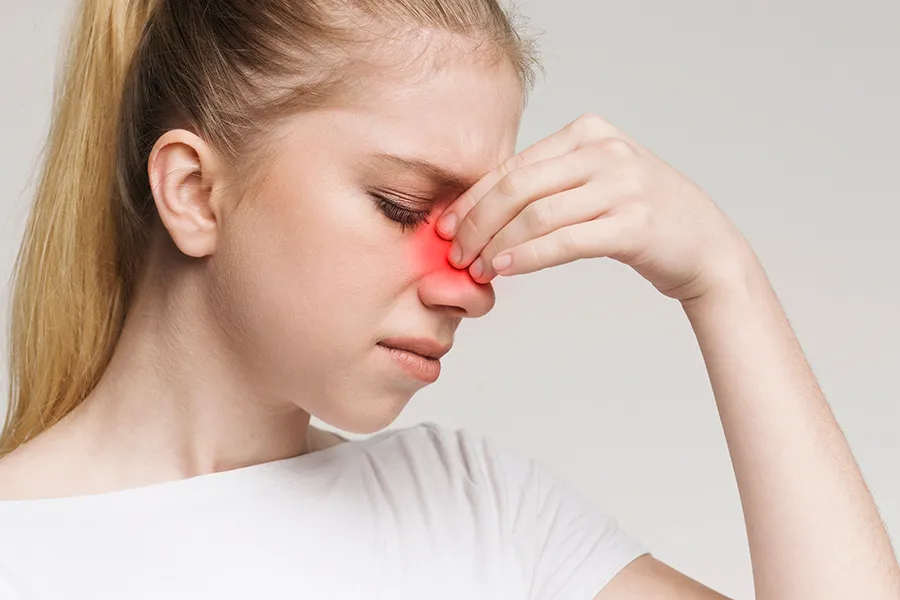 A woman grimaces and rubs the bridge of her nose due to sinus irritation. Get treatment for allergic rhinitis from Allen Green, M.D. of Center for Optimum Health in Los Angeles.