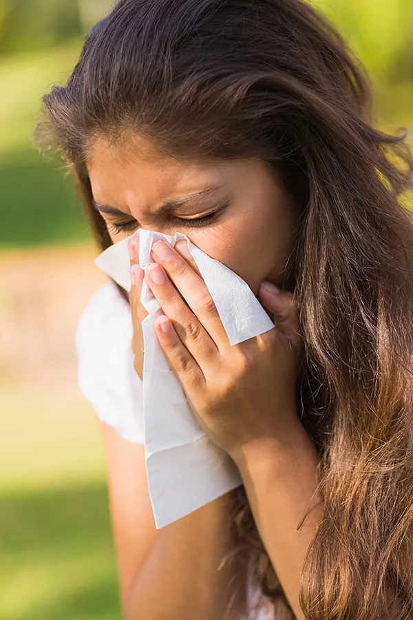 A woman with long brown hair sneezes into a handkerchief outdoors. Get allergy treatment services from Allen Green, M.D. of Center for Optimum Health in Los Angeles.