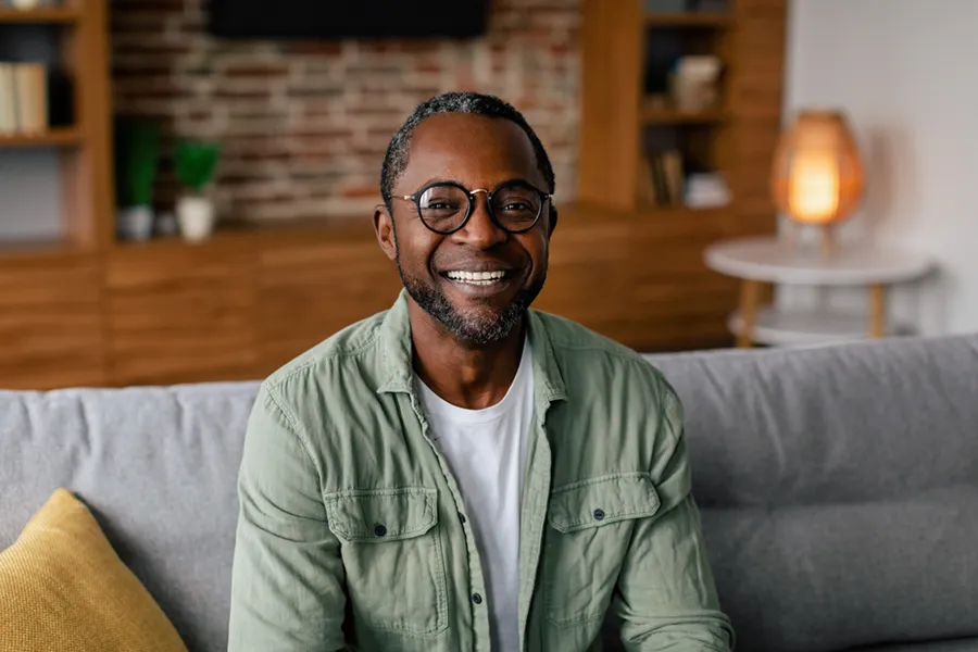 A middle aged black man in a green button up shirt sits on his couch smiling, happy with his treatment for andropause and low testosterone from Allen Green, M.D. of Center for Optimum Health in Los Angeles.
