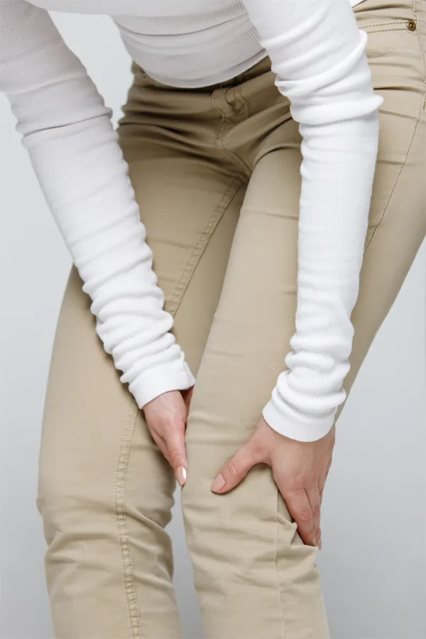 A woman wearing a white long-sleeve shirt and tan pants, clutching her knee, suffering from arthritis. Get arthritis treatment from Allen Green, M.D. of Center for Optimum Health in Los Angeles.