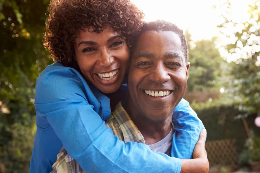 A middle-aged black couple outside, the woman embracing the man from behind pressing her cheek to his, both are smiling due to successful Autoimmune Disorder treatment from Allen Green, M.D. of Center for Optimum Health in Los Angeles.