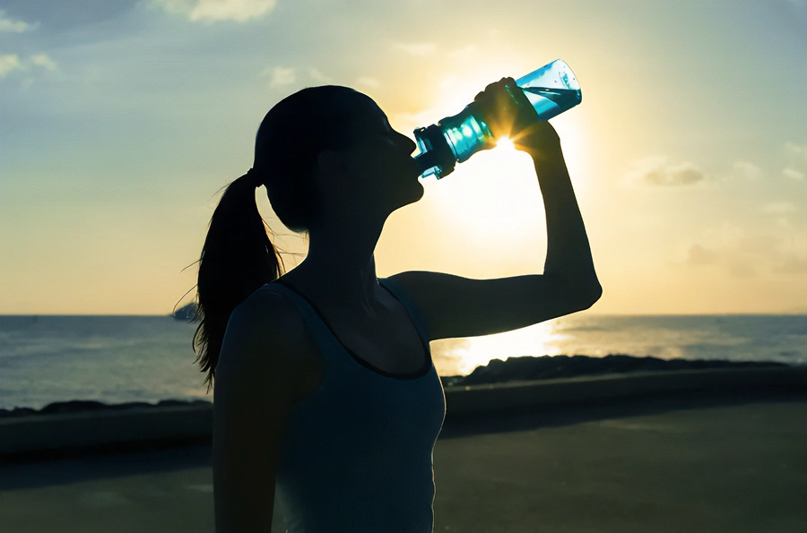 Chelation-and-Detoxification-Clinic A woman in exercise clothes drinking from a water bottle on the beach, with the water and sunset in the background. Get chelation and detoxification treatment from Allen Green, M.D. of Center for Optimum Health in Los Angeles.