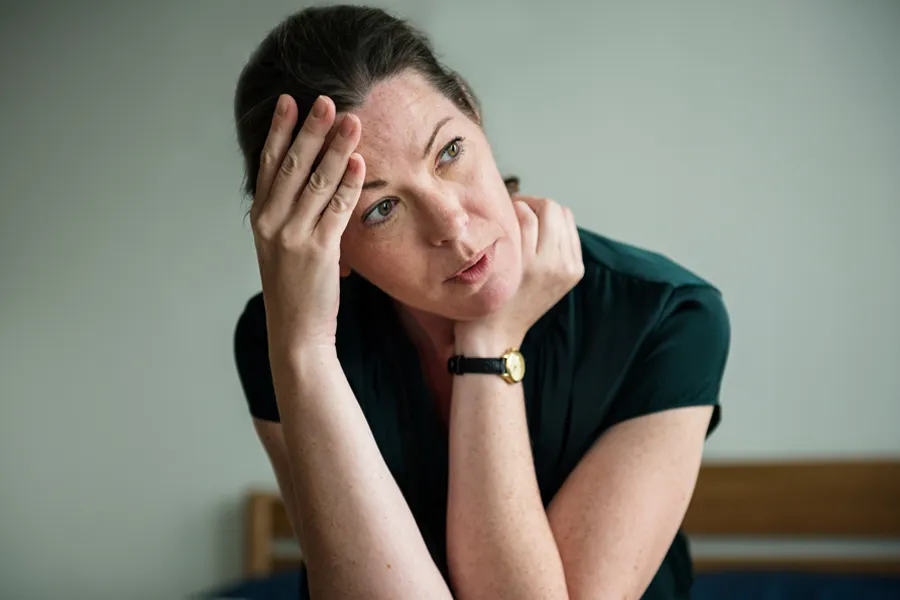 A woman in a dark blue dress sits on her bed holding her head in her hands with an exhausted expression. Get help for chronic fatigue syndrome from Allen Green, M.D. of Center for Optimum Health in [AERA].