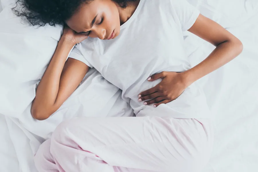 A dark-skinned woman in white clothes lays on her bed clutching her middle before getting treatment for Crohn's Disease from Allen Green, M.D. of Center for Optimum Health in Los Angeles.
