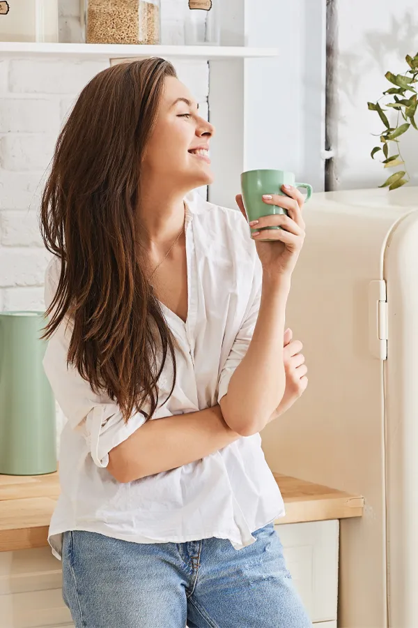 A woman with a white blouse an jeans drinking tea in her kitchen, smiling after getting treatment for Crohn's Disease from Allen Green, M.D. of Center for Optimum Health in Los Angeles.