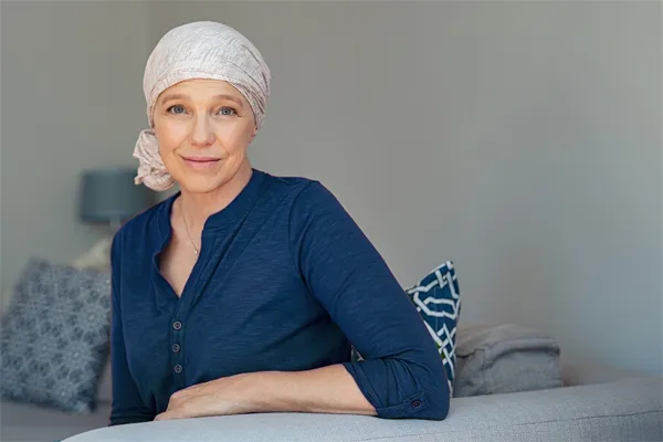 A woman wearing a blue shirt with a white scarf tied around her head sitting on a couch and smiling at the camera, representing sensitive functional oncology treatment from Allen Green, M.D. of Center for Optimum Health in Los Angeles.