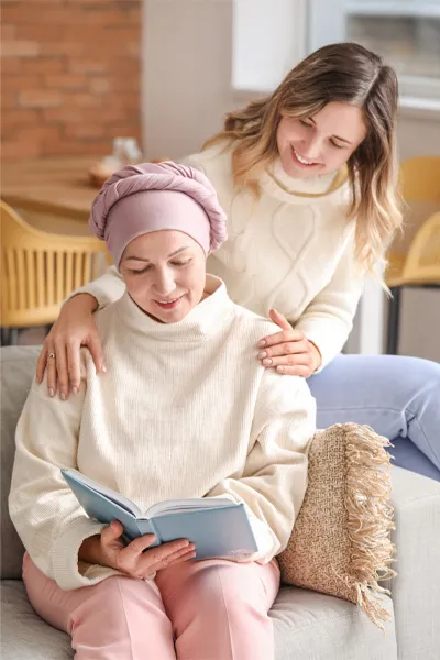 A woman with a scarf tied around her head sits on a couch reading while another woman sits behind her with her hands on her shoulders warmly, representing sensitive functional oncology treatment from Allen Green, M.D. of Center for Optimum Health in Los Angeles.