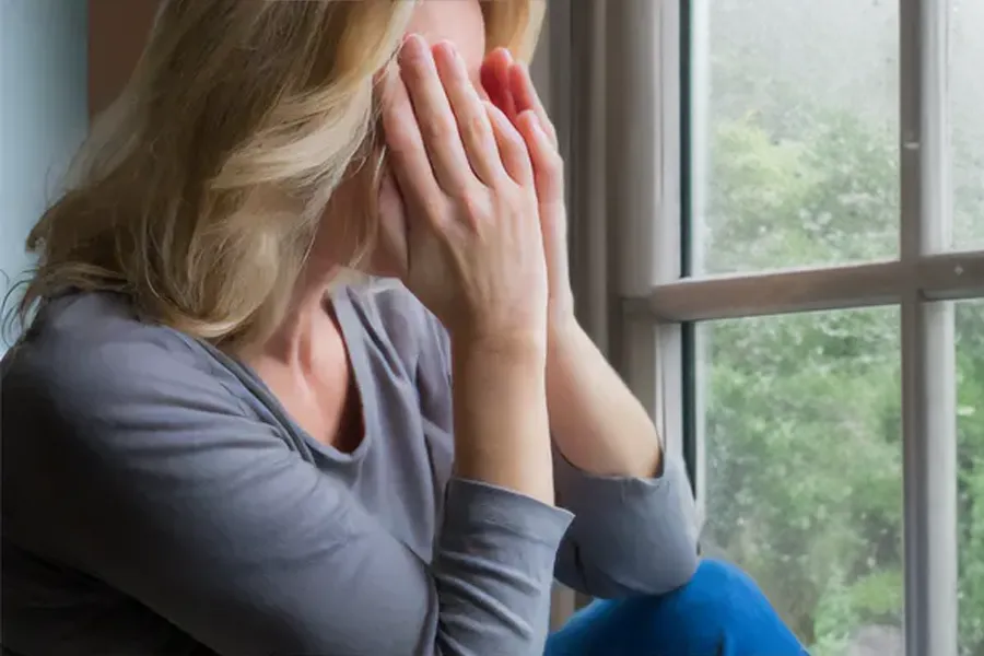 A blonde woman in a gray shirt sits in front of a window on a rainy day covering her eyes with her hands. Get treatment for Grave's Disease from Allen Green, M.D. of Center for Optimum Health in Los Angeles.