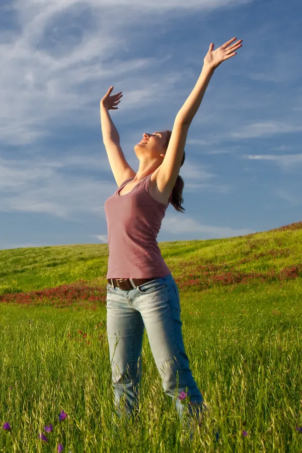 A woman in a tank top and jeans looks up with her arms outstretched to the sky in a field after successful treatment for Grave's Disease from Allen Green, M.D. of Center for Optimum Health in Los Angeles.