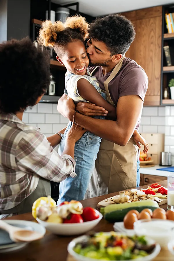A man holds his daughter and kisses her cheek as the family makes a healthy dinner in the kitchen. Get help for digestive issues and gut health from Allen Green, M.D. of Center for Optimum Health in Los Angeles.