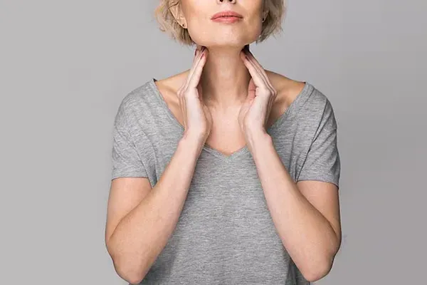 An older blonde woman in a gray t-shirt holds her hands to her neck checking for signs of thyroid problems. Get help for Hashimoto's Thyroiditis from Allen Green, M.D. of Center for Optimum Health in Los Angeles.