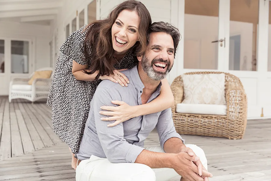 A middle-aged couple on a wooden porch with wicker furniture; the woman is playfully hugging the man from behind. They are benefiting from the health and wellness services from Allen Green, M.D. of Center for Optimum Health in Los Angeles.