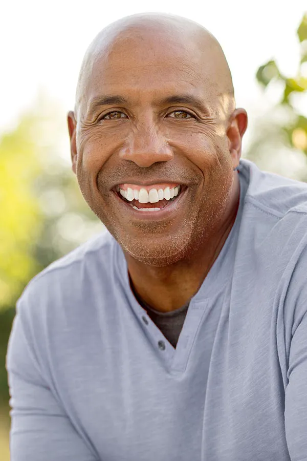A middle-aged black man in a gray v-neck t-shirt sits outside smiling, happy with the healthy aging peptide treatments from Allen Green, M.D. of Center for Optimum Health in Los Angeles
