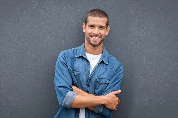 A man in a denim shirt stands smiling against a gray-blue wall, pleased with his testosterone hormone therapy from Allen Green, M.D. of Center for Optimum Health in Los Angeles.