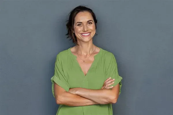 A woman in a green shirt stands smiling against a gray-blue wall, pleased with her hormone therapy from Allen Green, M.D. of Center for Optimum Health in Los Angeles.