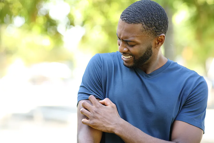 A black man in a blue shirt scratches his arm while outside. Get treatment for insect allergies from Allen Green, M.D. of Center for Optimum Health in Los Angeles.
