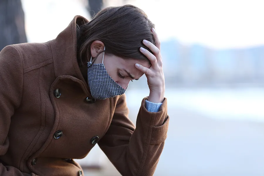 A woman in a brown coat and gray-blue mask sits outside holding her head in pain from long covid. Get treatment for long covid from Allen Green, M.D. of Center for Optimum Health in Los Angeles.