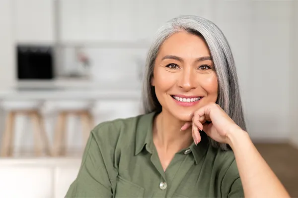An asian woman with long gray hair and a green shirt sitting and smiling on her couch, happy with her menopause care from [NAME} in Los Angeles.