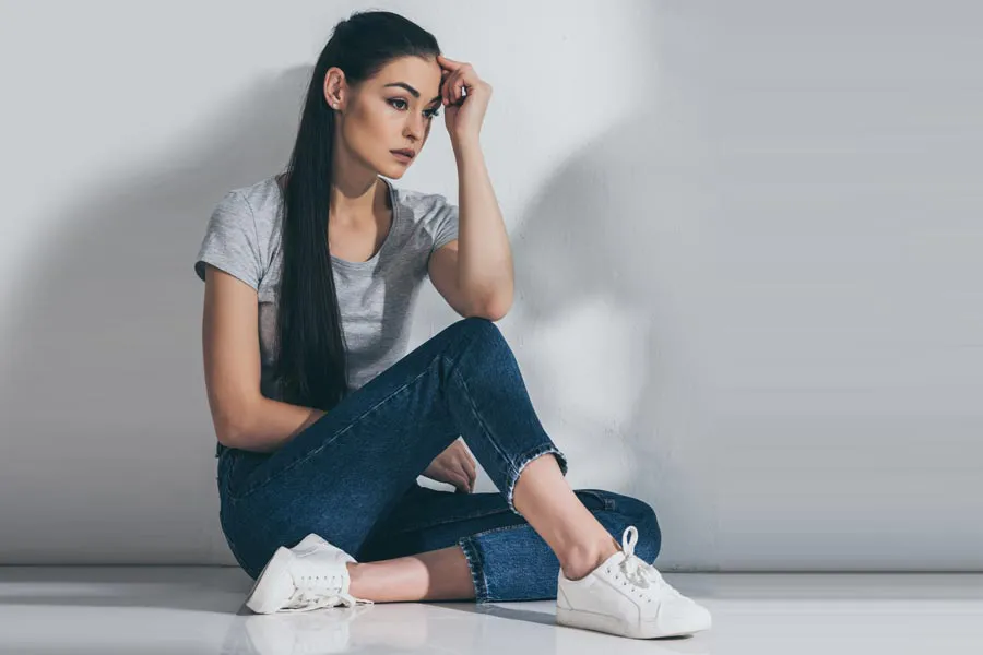A young brunette woman in a gray t-shirt and jeans sits against a gray wall with her head resting on her hand and a distressed expression. Get help for Polycystic Ovarian Syndrome (PCOS) from Allen Green, M.D. of Center for Optimum Health in Los Angeles.