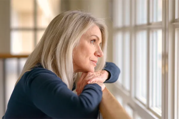 A middle-aged woman with with a somber expression stares out the window. Get treatment for Premenstrual Dysphoric Disorder from Allen Green, M.D. of Center for Optimum Health in Los Angeles.