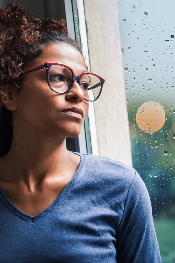 A black woman with with a somber expression and glasses and a gray shirt stares out the window on a rainy day. Get treatment for Premenstrual Dysphoric Disorder from Allen Green, M.D. of Center for Optimum Health in Los Angeles.