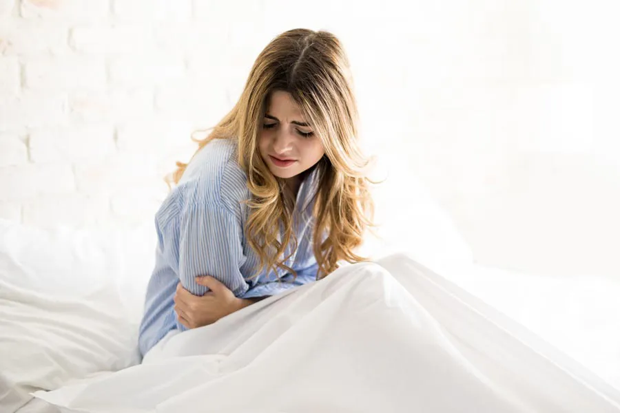 PMS-Therapy A woman with long blonde hair sits in her bed holding her stomach in pain, considering getting care for PMS from Allen Green, M.D. of Center for Optimum Health in Los Angeles.