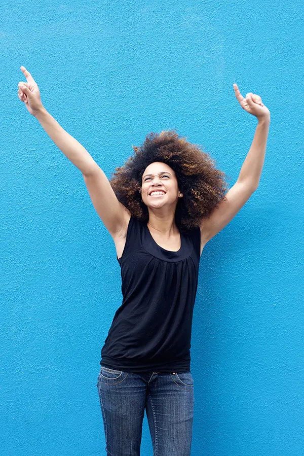 PMS-Treatment A woman in a dark blue tank top standing in front of a bright blue wall, raising her arms in celebration of relief from PMS from Allen Green, M.D. of Center for Optimum Health in Los Angeles.
