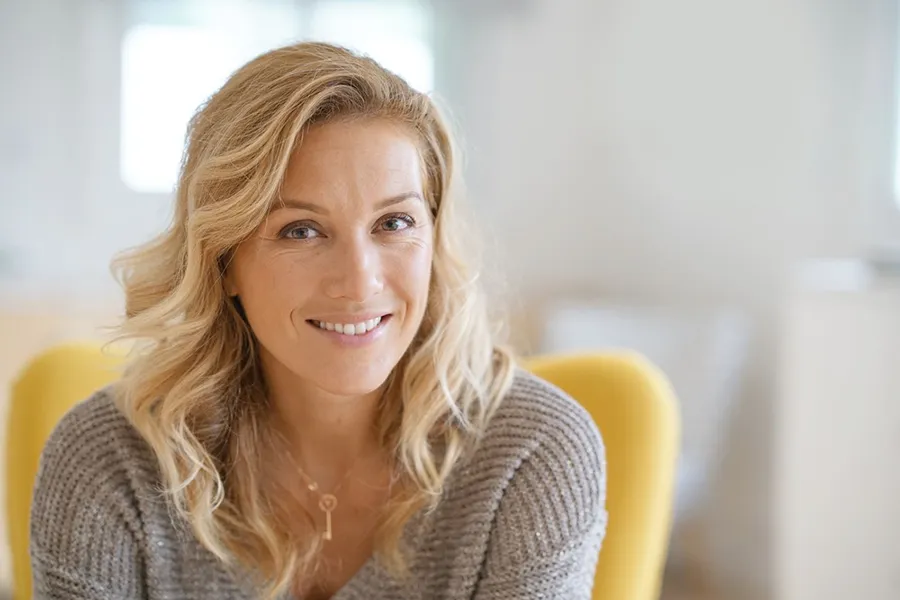 Perimenopause-Doctor A blonde woman wearing a a gray sweater sits inside in a yellow chair, smiling and happy with her perimenopause treatment from Allen Green, M.D. of Center for Optimum Health in Los Angeles.