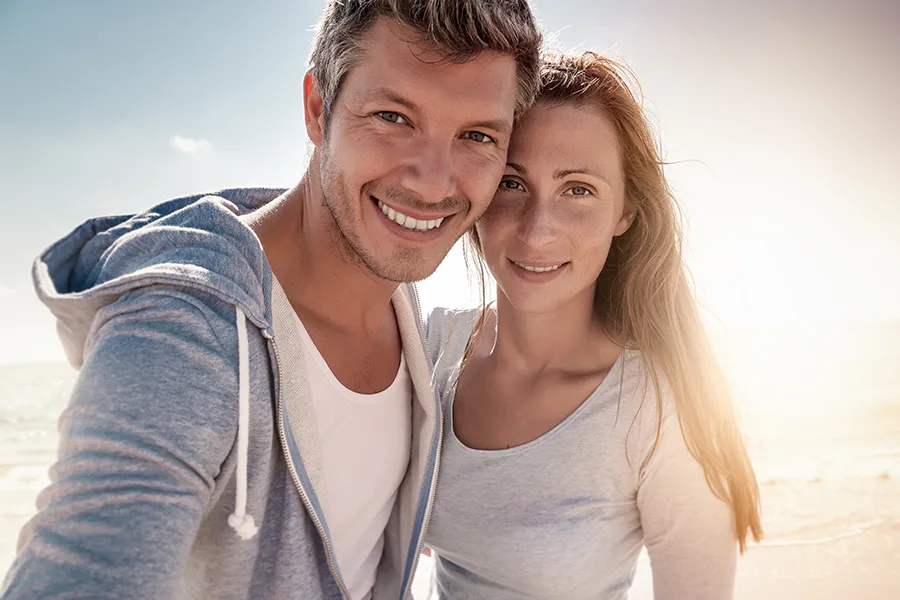 A middle-aged couple stands on the beach with the sun behind them. Get treatment for medical skin conditions from Allen Green, M.D. of Center for Optimum Health in Los Angeles.