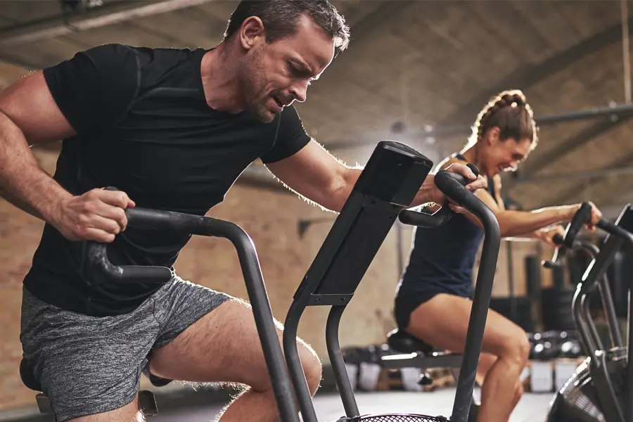 A couple in a brick gym work as hard as they can on exercise bikes, inspired by successful Tirzepatide treatment from Allen Green, M.D. of Center for Optimum Health in Los Angeles.