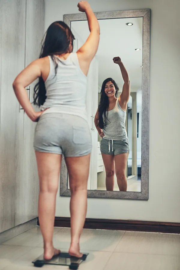 A woman in workout clothes stands on a scale looking in the mirror and pumping her hands in celebration of her successful Tirzepatide treatment from Allen Green, M.D. of Center for Optimum Health in Los Angeles.