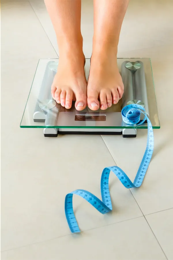 Weight-Loss-Resistance-Treatment Close-up of a woman's feet standing on a scale, with measuring tape by her toes, getting treatment for weight loss resistance from Allen Green, M.D. of Center for Optimum Health in Los Angeles.